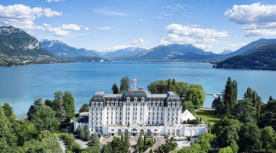 Vue du palais impérial de la Ville d'Annecy en face du lac, entouré de quelques arbres et montagnes en arrière-plan